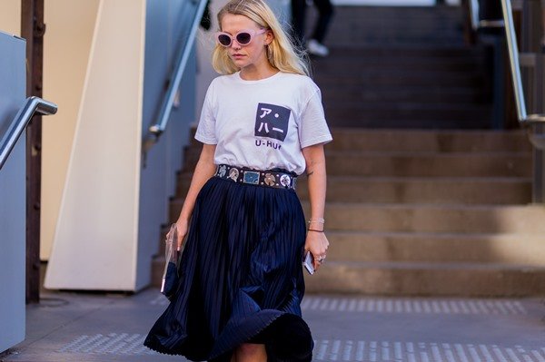 Mulher branca e com cabelo liso louro, andando pela rua, com óculos escuros de armação lilás. Ela usa uma camiseta branca, com um desenho de símbolos japoneses pretos, uma saia plissada preta e um cinto de taxas.